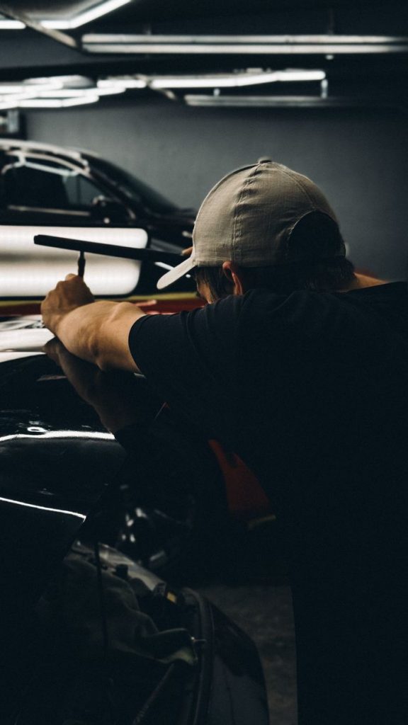 A mechanic wearing a cap examines a car under low light in a garage.
