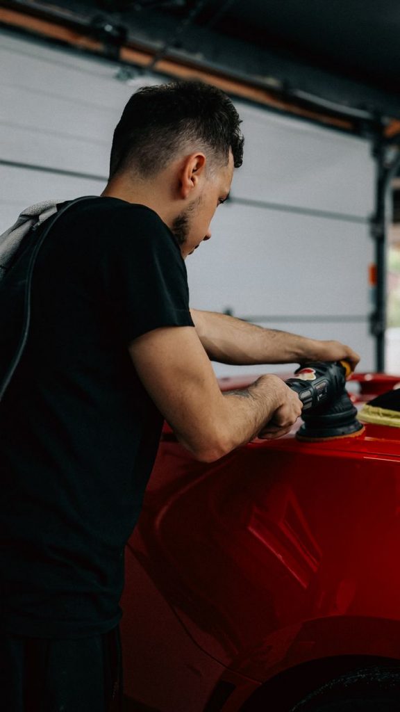 Young man using a polisher on a shiny red car in a garage setting.